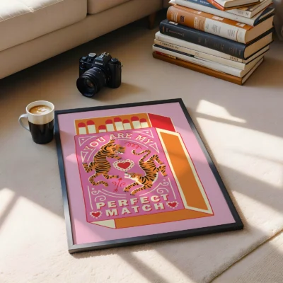 A framed artwork displaying two tigers entwined around a heart with the text 'You Are My Perfect Match', accompanied by a coffee cup and stack of books on a rug.