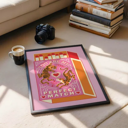 A framed artwork displaying two tigers entwined around a heart with the text 'You Are My Perfect Match', accompanied by a coffee cup and stack of books on a rug.