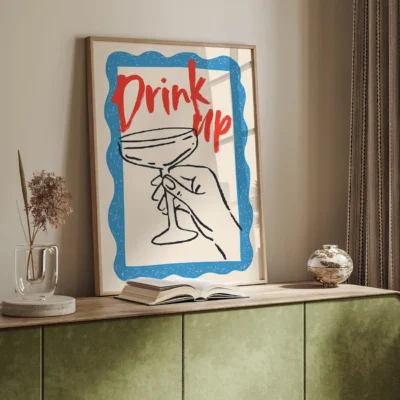 A framed artwork displaying a hand holding a cocktail glass with the text 'Drink up' in red on a blue background